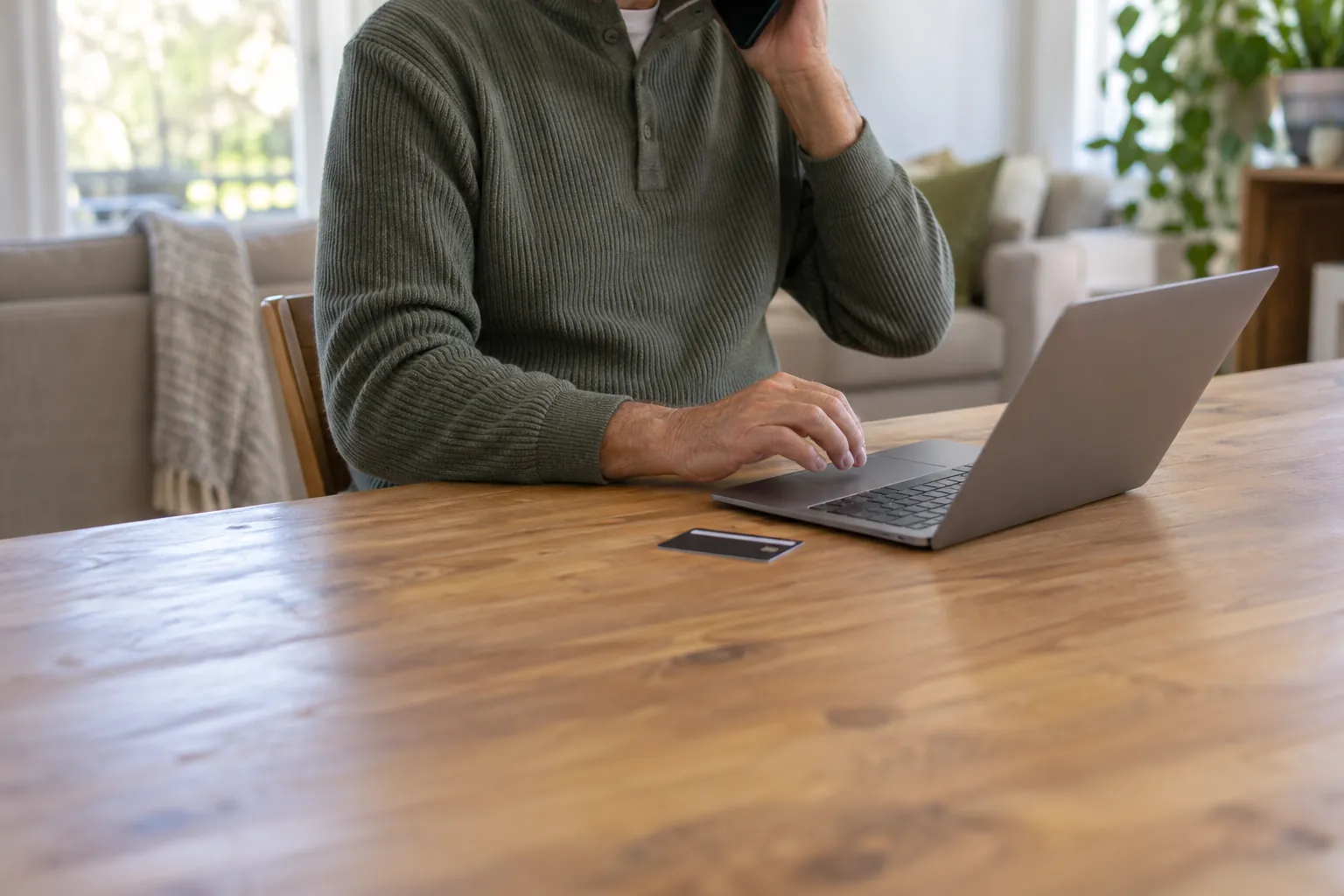 Un uomo al telefono davanti a un portatile aperto, con la carta bancaria appoggiata sul tavolo